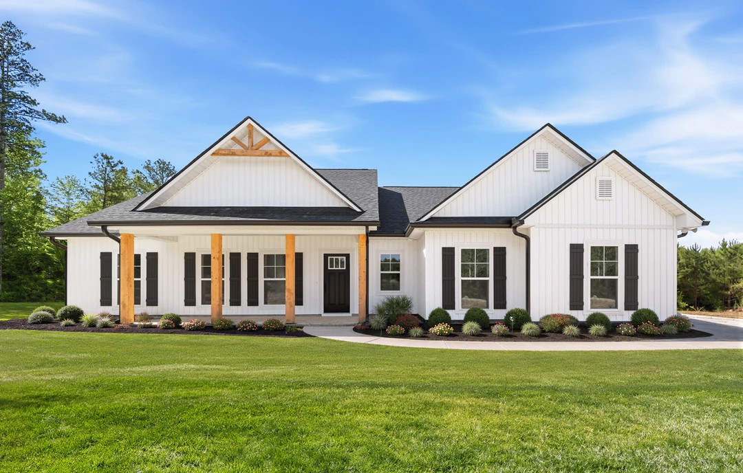 White farmhouse-style home with black front door, white siding, and a covered porch, set on a green lawn with flower beds, a walkway, and mature trees under a blue sky.