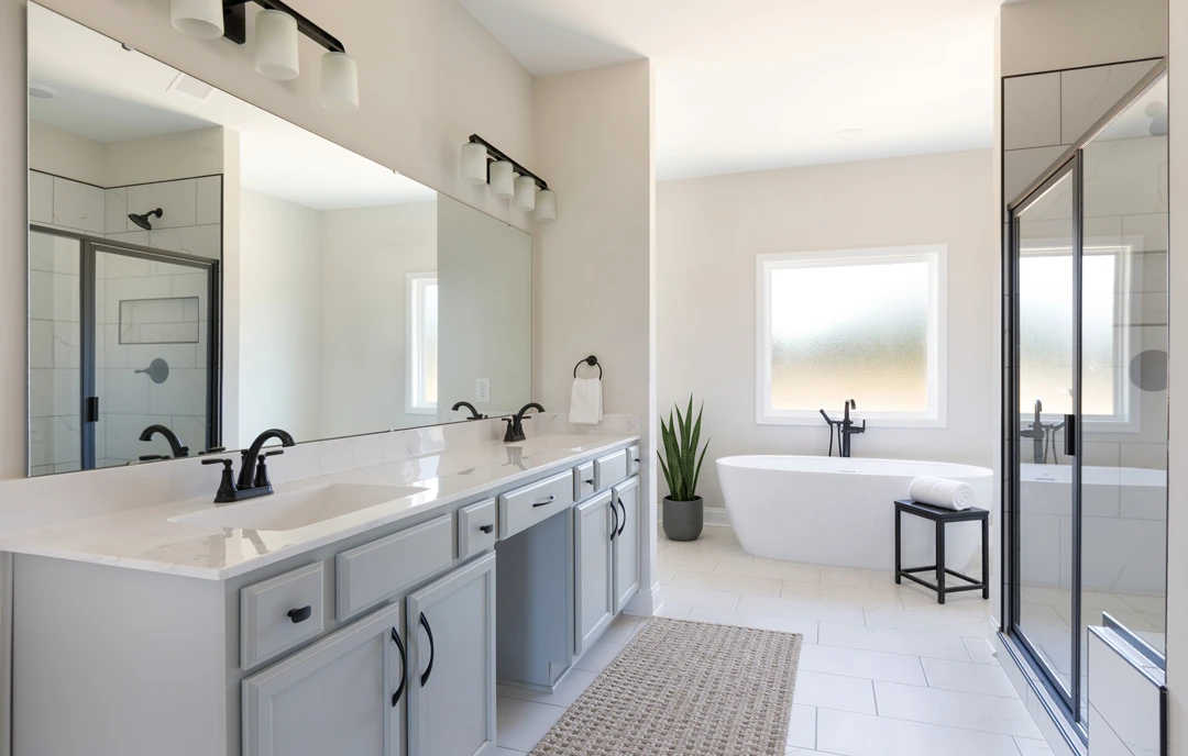Modern bathroom with a freestanding white tub beside a black side table holding a rolled white towel, a potted plant, a glass shower door, and a light tile floor with a small rug.