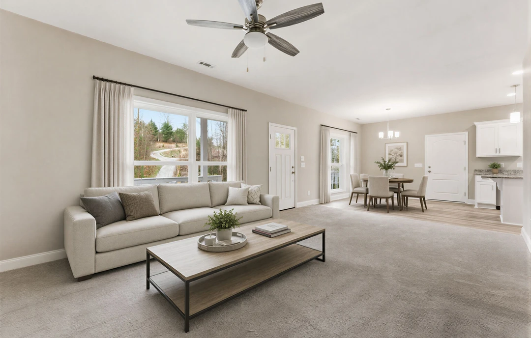 Living room with a gray couch topped with pillows, a coffee table holding a potted plant, ceiling fan with light, and a window showing a view of the road outside.