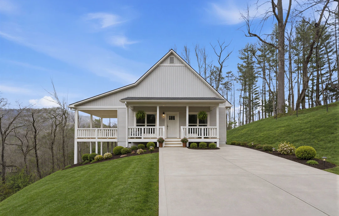 Two-story farmhouse with light siding, covered front porch, and a paved driveway bordered by green lawn, bushes, and mature trees under a partly cloudy sky.