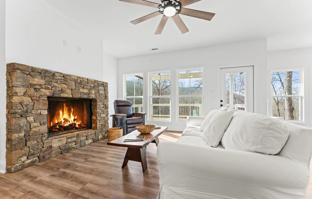 Living room with wood flooring, a stone fireplace with fire burning, a ceiling fan with light, and a couch with white pillows; a table with a basket sits in front of the fireplace.