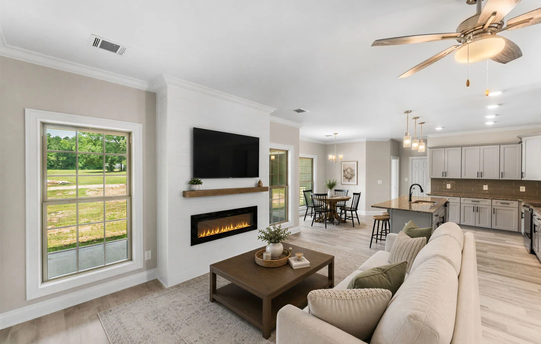 Living room with a gray couch facing a fireplace and wall-mounted TV, coffee table with a plant in front, large window showing a grassy field, and ceiling fan overhead.