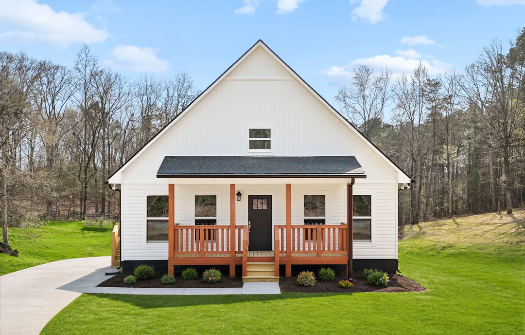 White farmhouse-style house with a wooden front porch, black door, and large windows, set on a green lawn with trees and cloudy sky in the background.