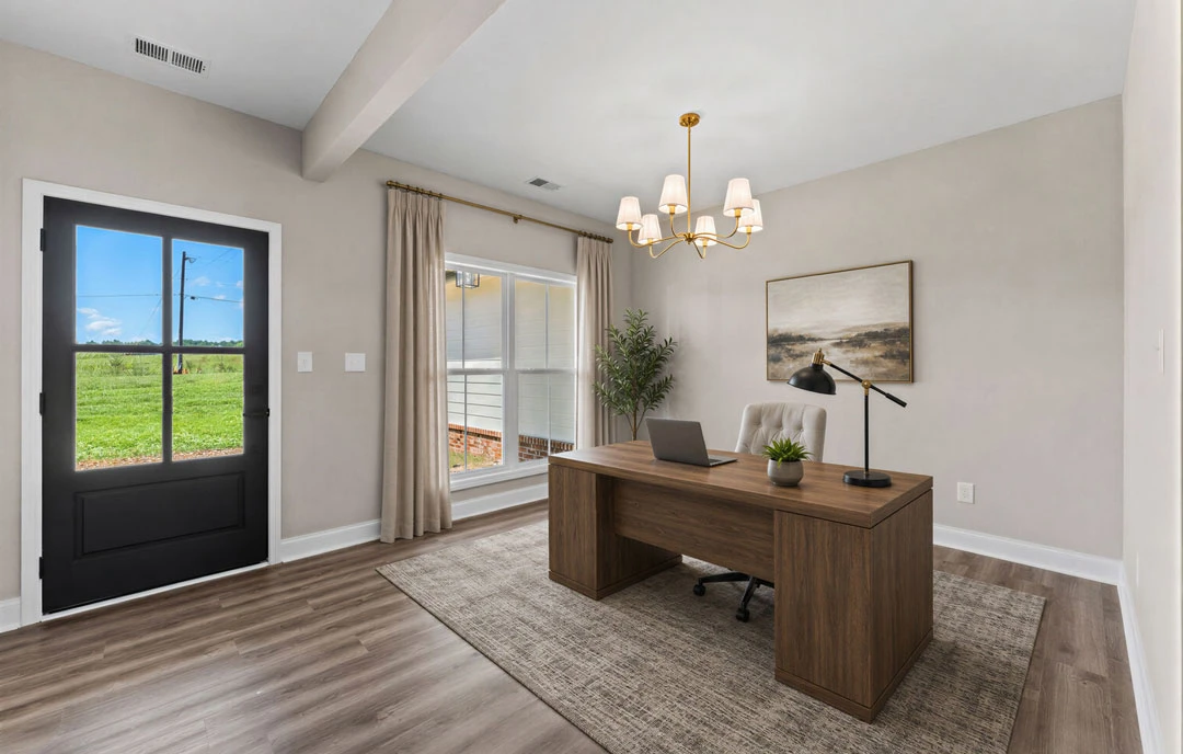 Den with wood flooring, a desk holding a laptop and plant, a lamp, and a painting on the wall; window and door offer views of a house and field.