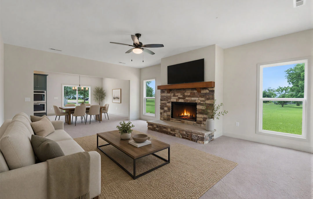 Living room with a stone fireplace, gray couch draped with a blanket, coffee table holding a bowl, ceiling fan with light, and window overlooking a grassy field; dining area