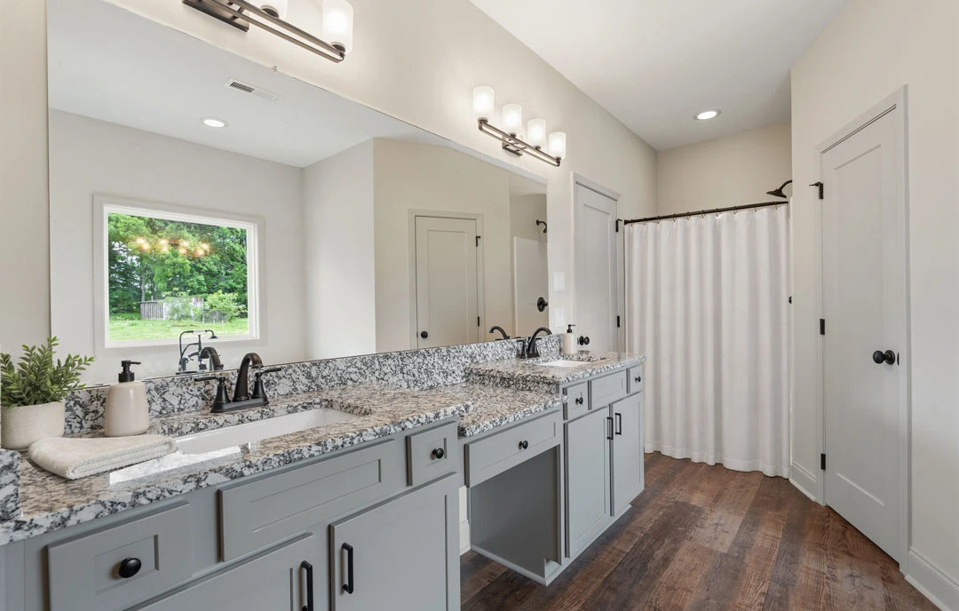 Bathroom with granite countertops, white cabinetry, a shower with a white curtain, and a potted plant near a window showing trees outside.