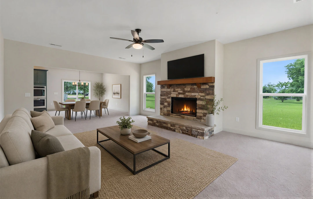 Living room with a stone fireplace, gray couch draped with a blanket, coffee table holding a bowl, ceiling fan with light, and large window overlooking a green field; dining area