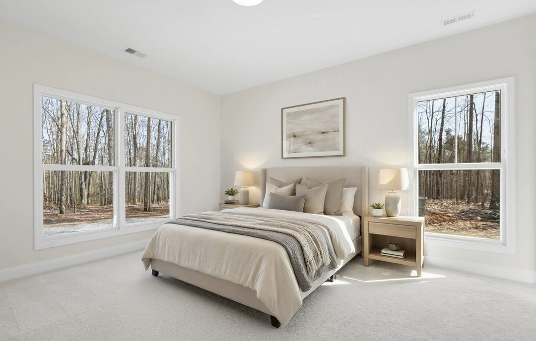 Bedroom with a white bed frame, white bedding, a nightstand, a shelf holding books and a bowl, framed artwork on the wall, and large windows showing trees outside.