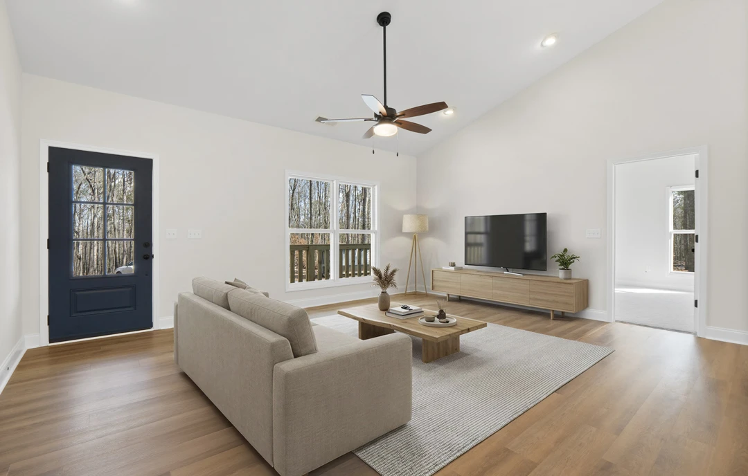 Living room with a gray couch facing a wall-mounted television, wood or laminate flooring, a coffee table with books and a vase, and a ceiling fan overhead; a door with a window