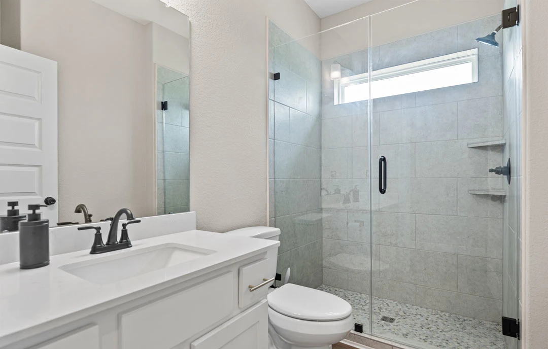 Bathroom with a glass-enclosed shower, white toilet, grey container with black lid, close-up of a sink and faucet, wall-mounted light, and stone countertops.