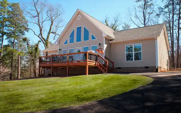 Single-story house with large windows, a wooden deck with stairs, and a grassy lawn surrounded by trees under a clear sky.