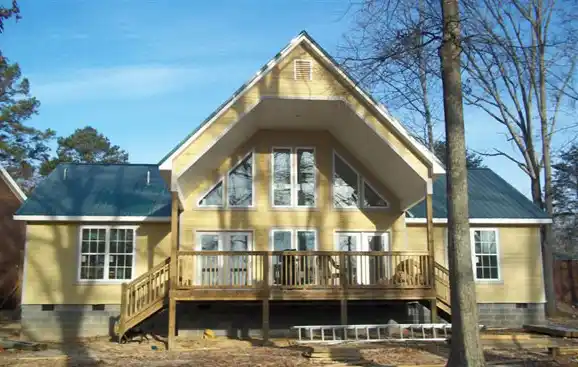 Yellow house with a green roof, large windows, a wooden deck with railing, and several trees in the yard.