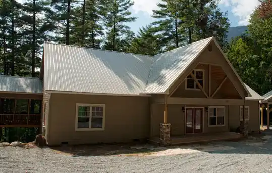 Single-story house with a metal roof, multiple windows, and a covered porch, surrounded by tall trees and a gravel driveway.