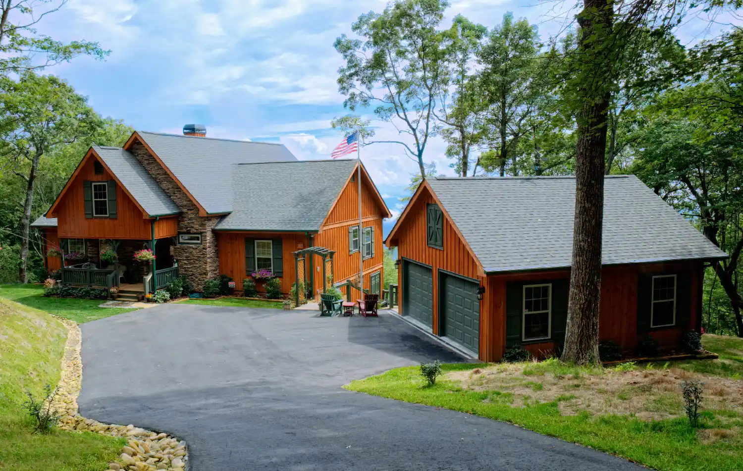 A house with wood siding and attached garages sits among trees, with a paved driveway, grassy areas, and an American flag visible.
