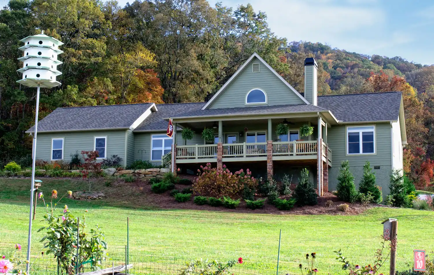 Single-story house with a large covered porch, fenced yard, landscaped garden, birdhouse pole, and surrounding trees on a grassy lot.