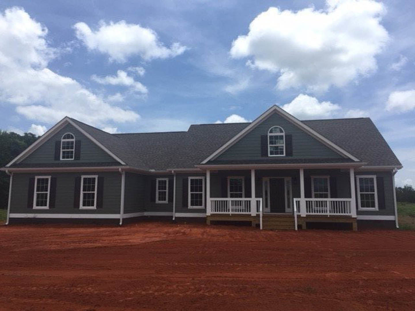 Two-story house under construction with white balcony railing, covered porch, red dirt road and field with tire tracks, blue sky and clouds overhead, white-framed windows