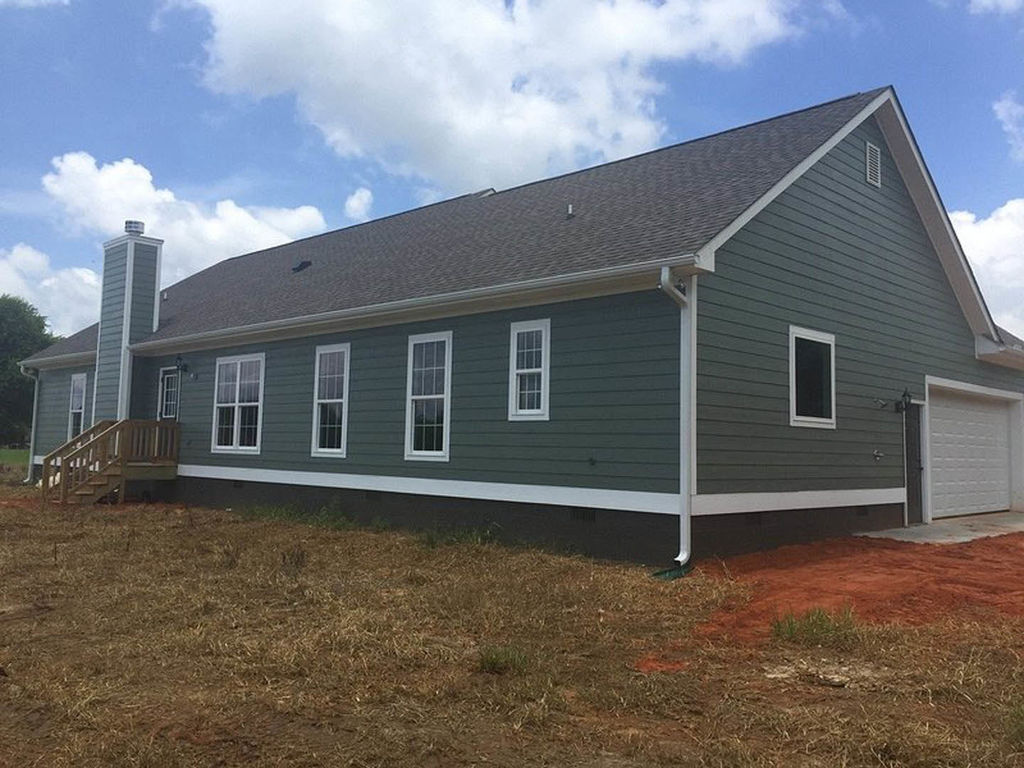 Two-story home with light siding, white-framed windows, wooden exterior staircase, fenced grassy yard, and blue sky with clouds