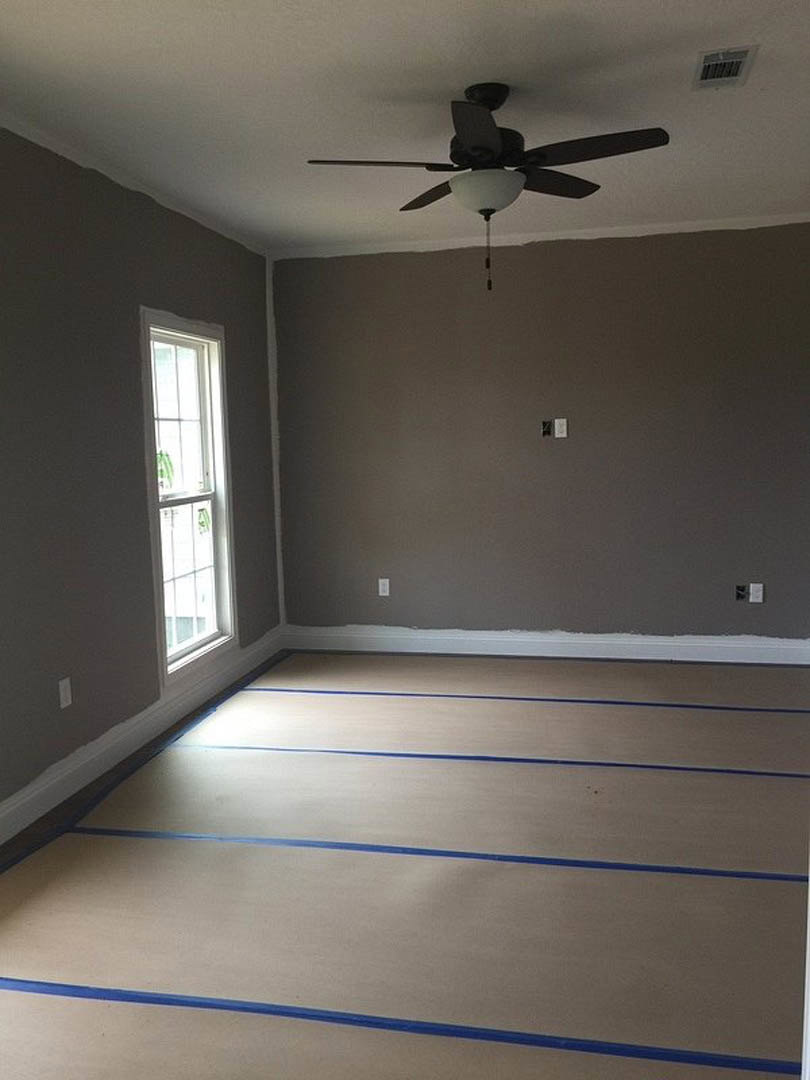 Ceiling fan with light fixture, white ceiling vent, window with white frame, blue painter’s tape on light-colored flooring, smooth plaster walls