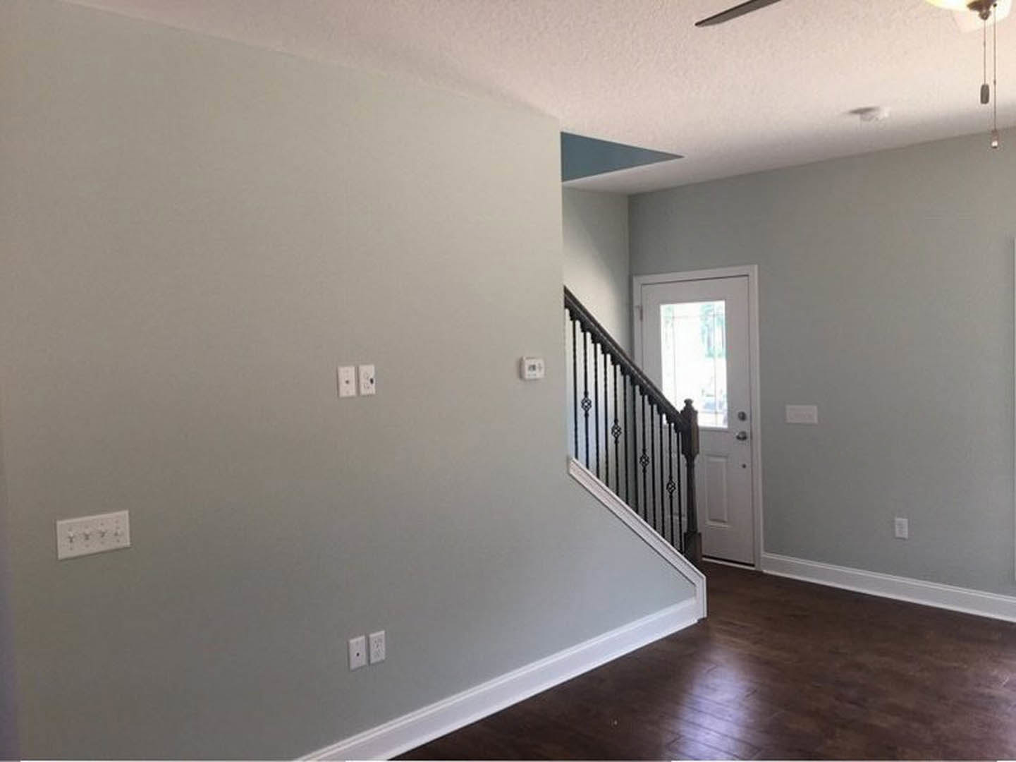 Hallway with white plaster walls, hardwood laminate flooring, wooden staircase with handrail, white door, and visible light switches and outlets