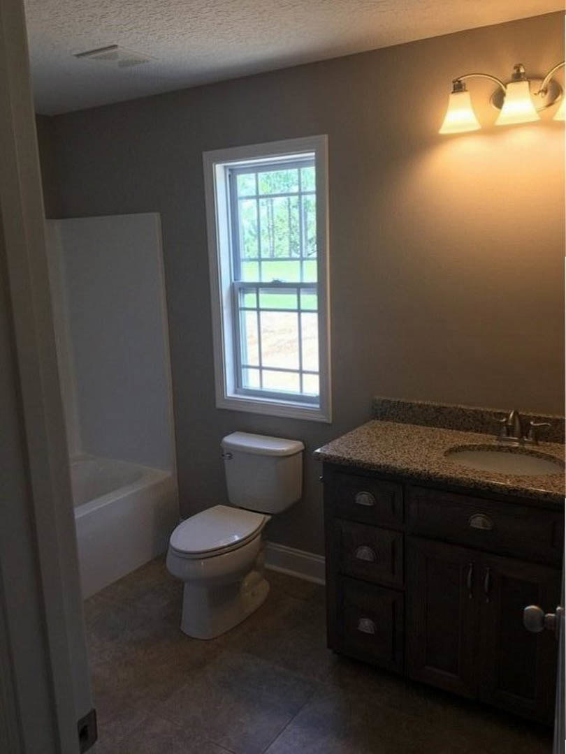 White porcelain toilet with closed seat, rectangular sink with chrome faucet, wall-mounted mirror above sink, two-bulb light fixture, tiled walls, window with exterior security