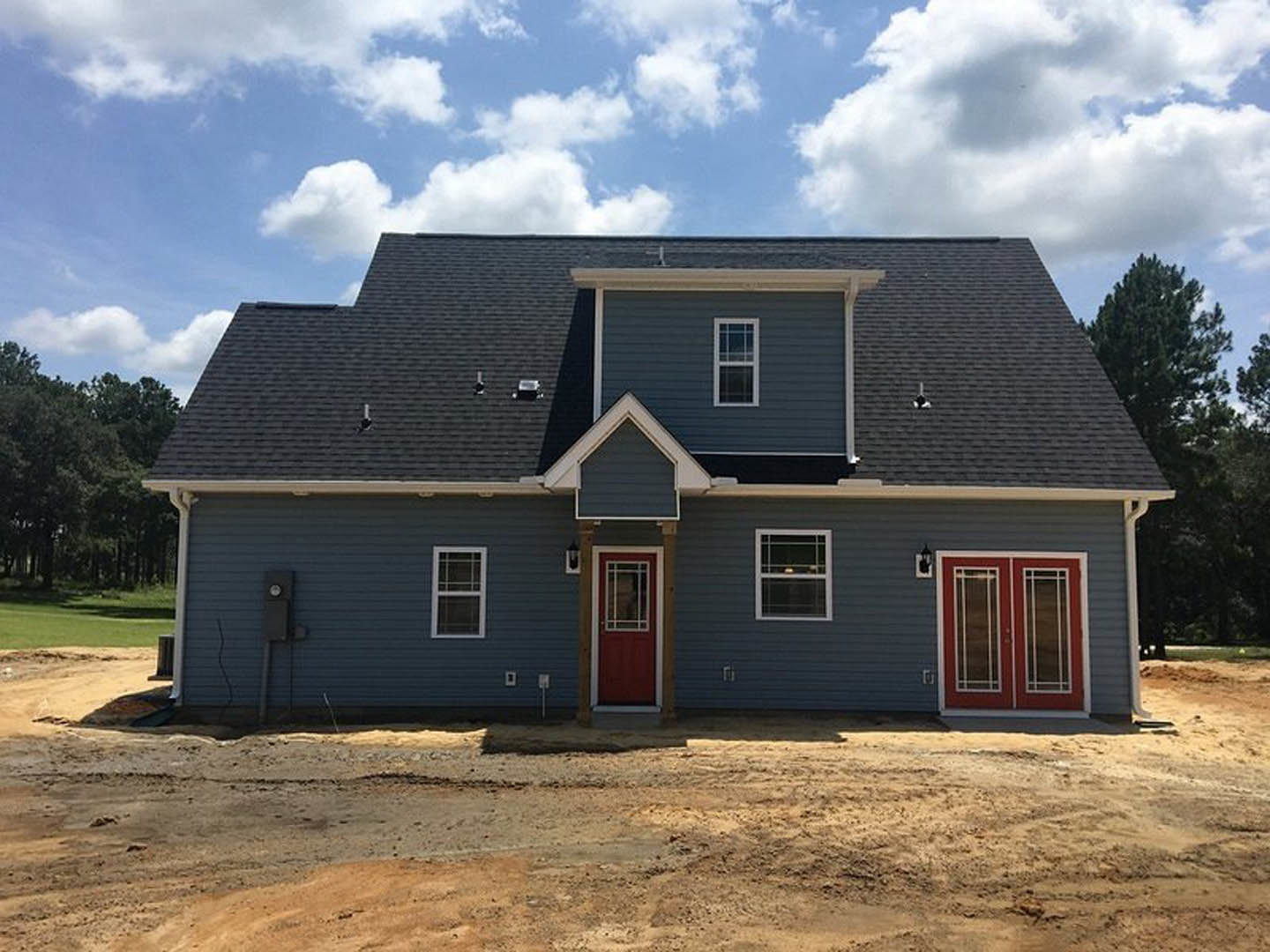 White cottage-style home with red double front door featuring glass panes, white trim, front window with candle, gray shingled roof, cloudy sky, and trees in the background