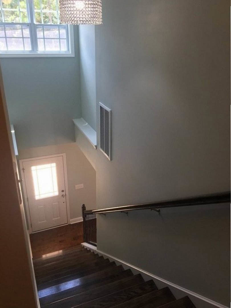 Wood staircase with metal handrail leading to a white door with glass window, tree visible through window, plaster walls, vent near baseboard, light-colored flooring