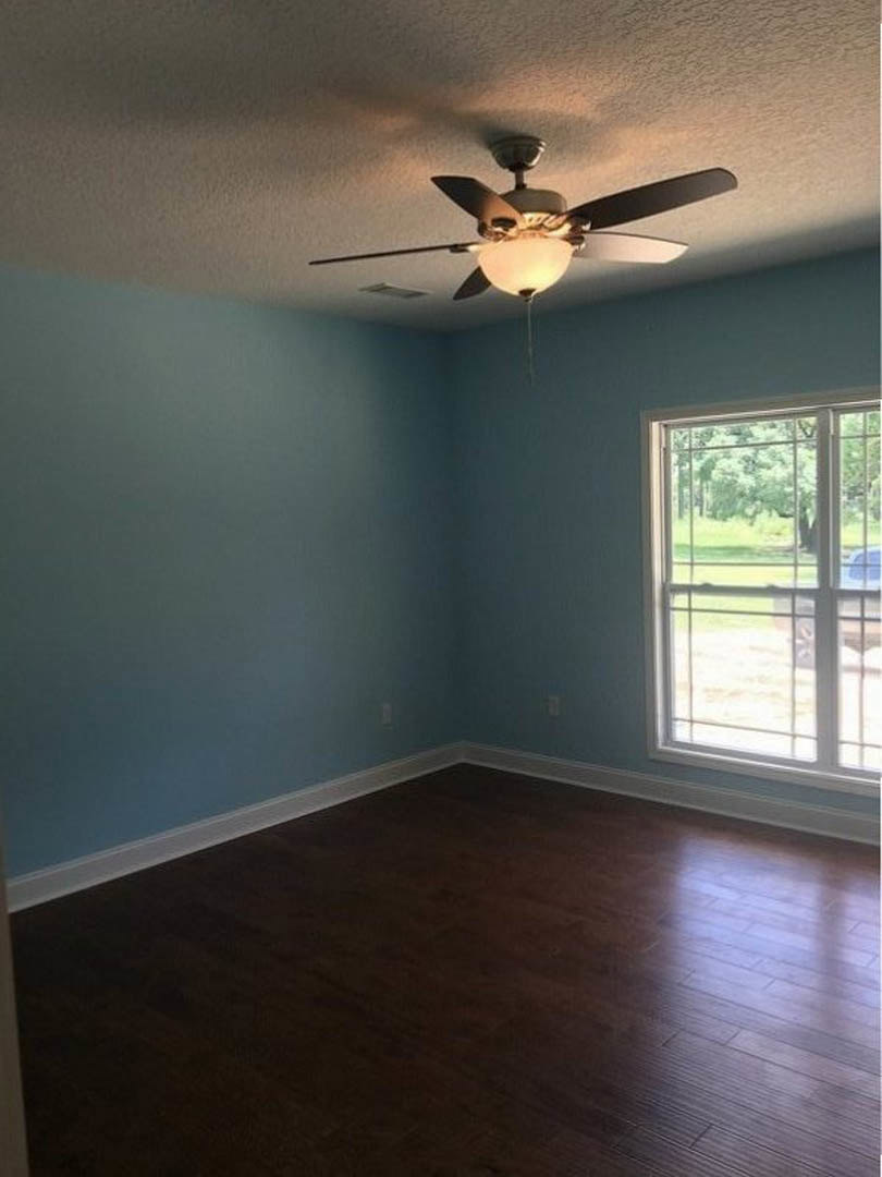 Ceiling fan with light fixture mounted on white plaster ceiling, dark hardwood floor with white baseboard trim, large window overlooking grassy yard and trees