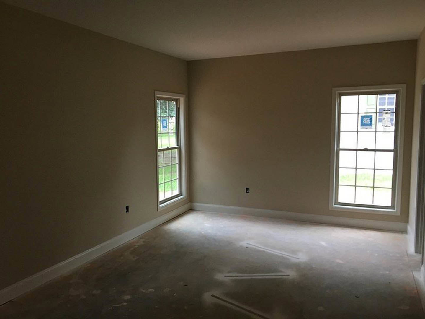 Sunlit room with concrete flooring, white plaster walls, large window overlooking grassy yard, blue sign with white text on window