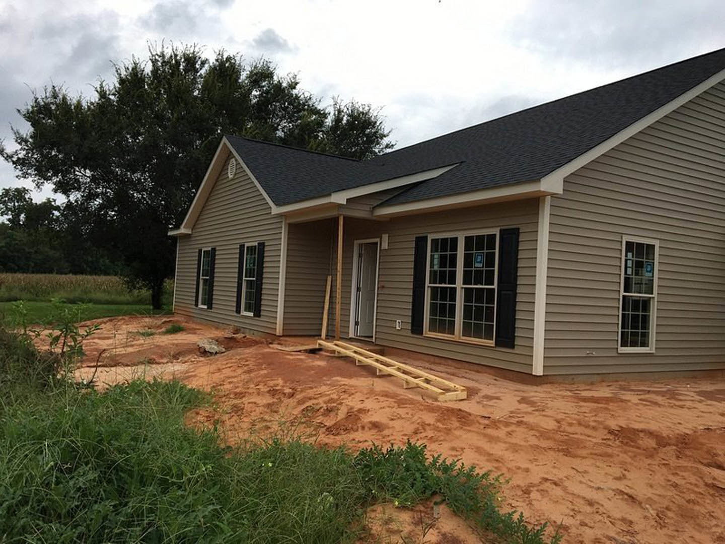 Partially built house with exposed wooden framing, multi-pane windows, white metal-framed door, scattered beams on ground, surrounded by grassy field and a few trees under cloudy