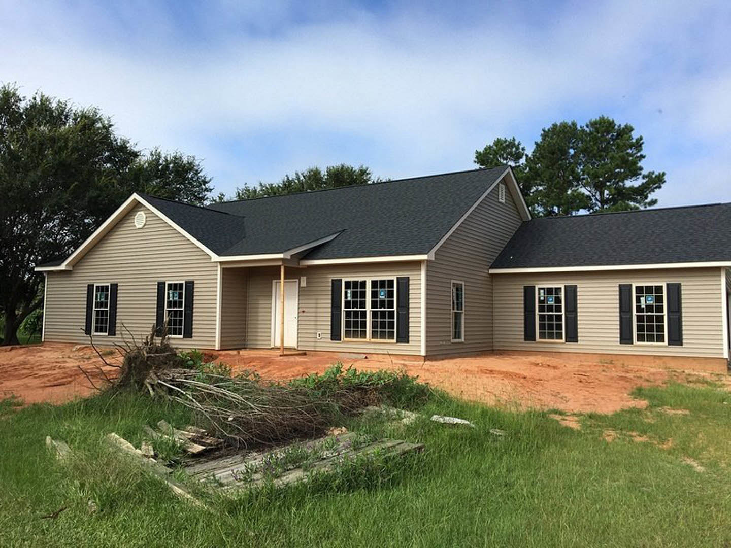 Two-story house under construction with black roof, white-framed windows, unfinished exterior walls, grassy yard, and mature trees in background
