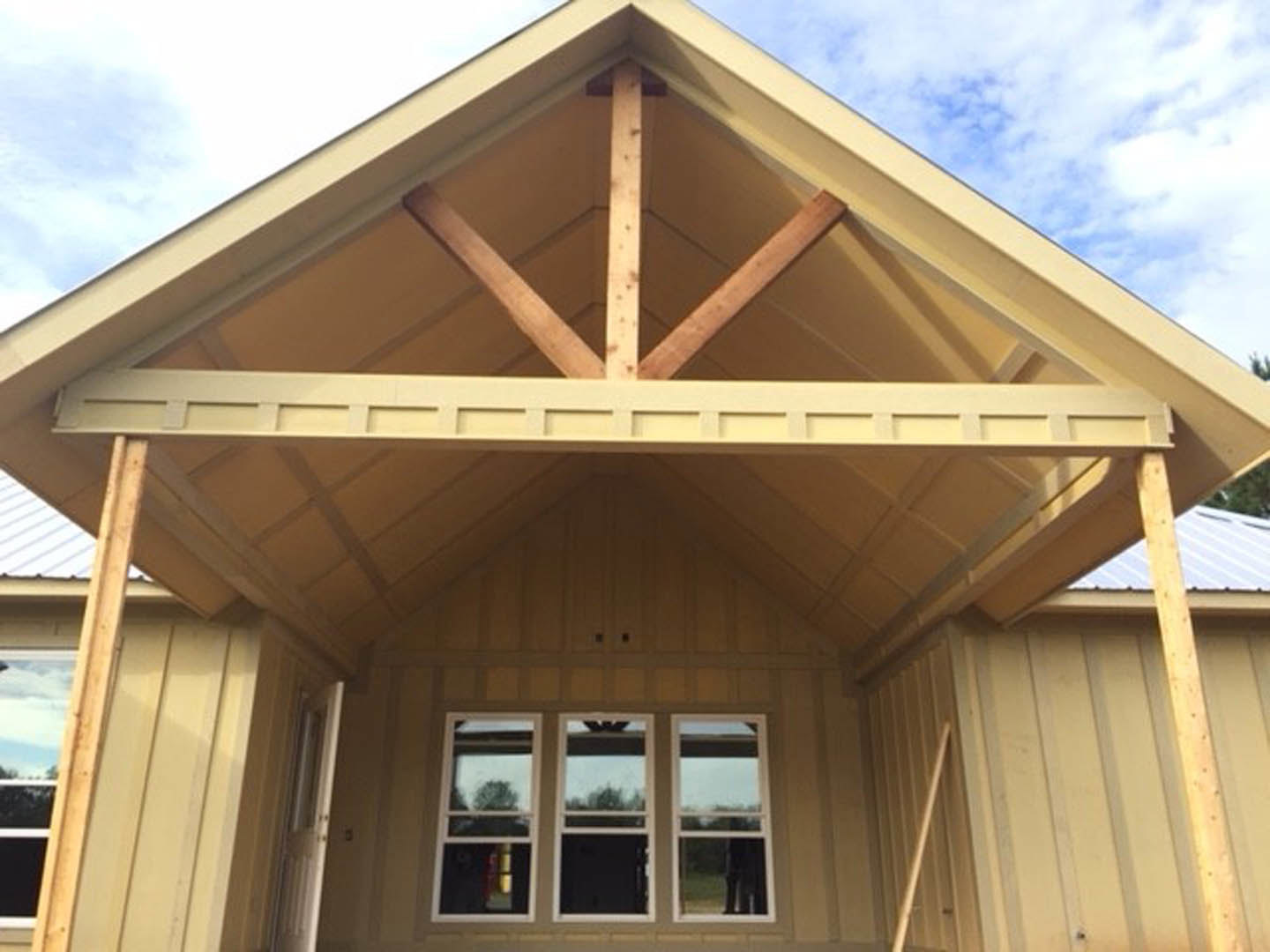 Two-story house under construction with gray siding, black front door, row of white-framed windows, exposed wooden beams, and trees visible through windows