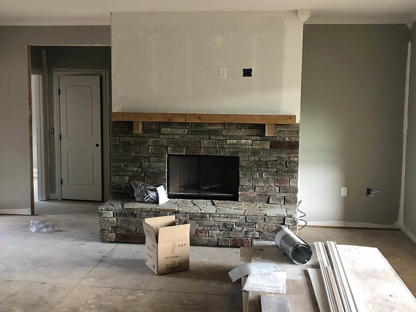 Stone fireplace with white mantel in living room, open cardboard box on hearth, white door with black handle, brick accent wall, hardwood floors, plaster ceiling