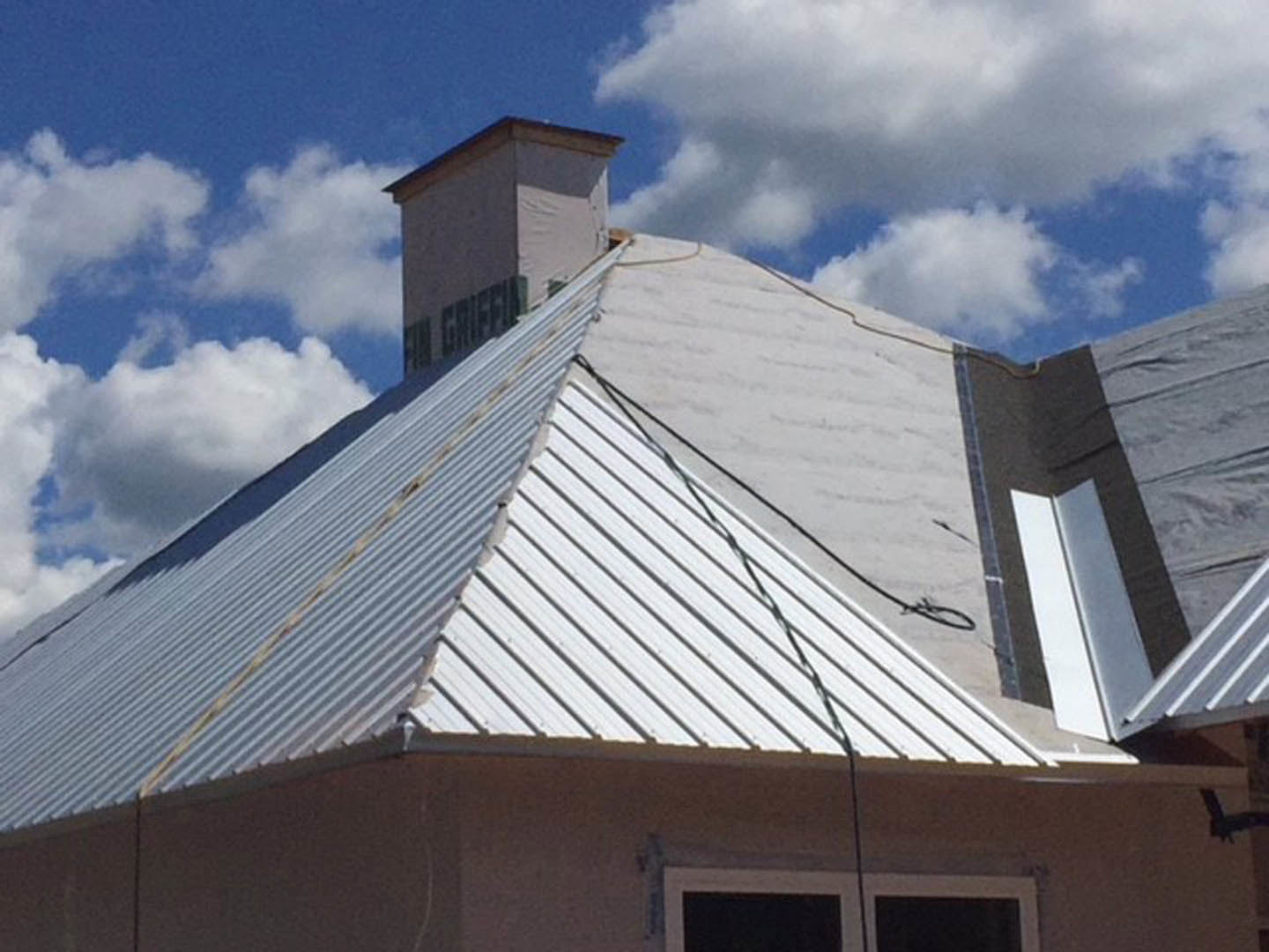 Gray shingle roof with brick chimney, partially constructed, adjacent to white-framed window, under cloudy sky