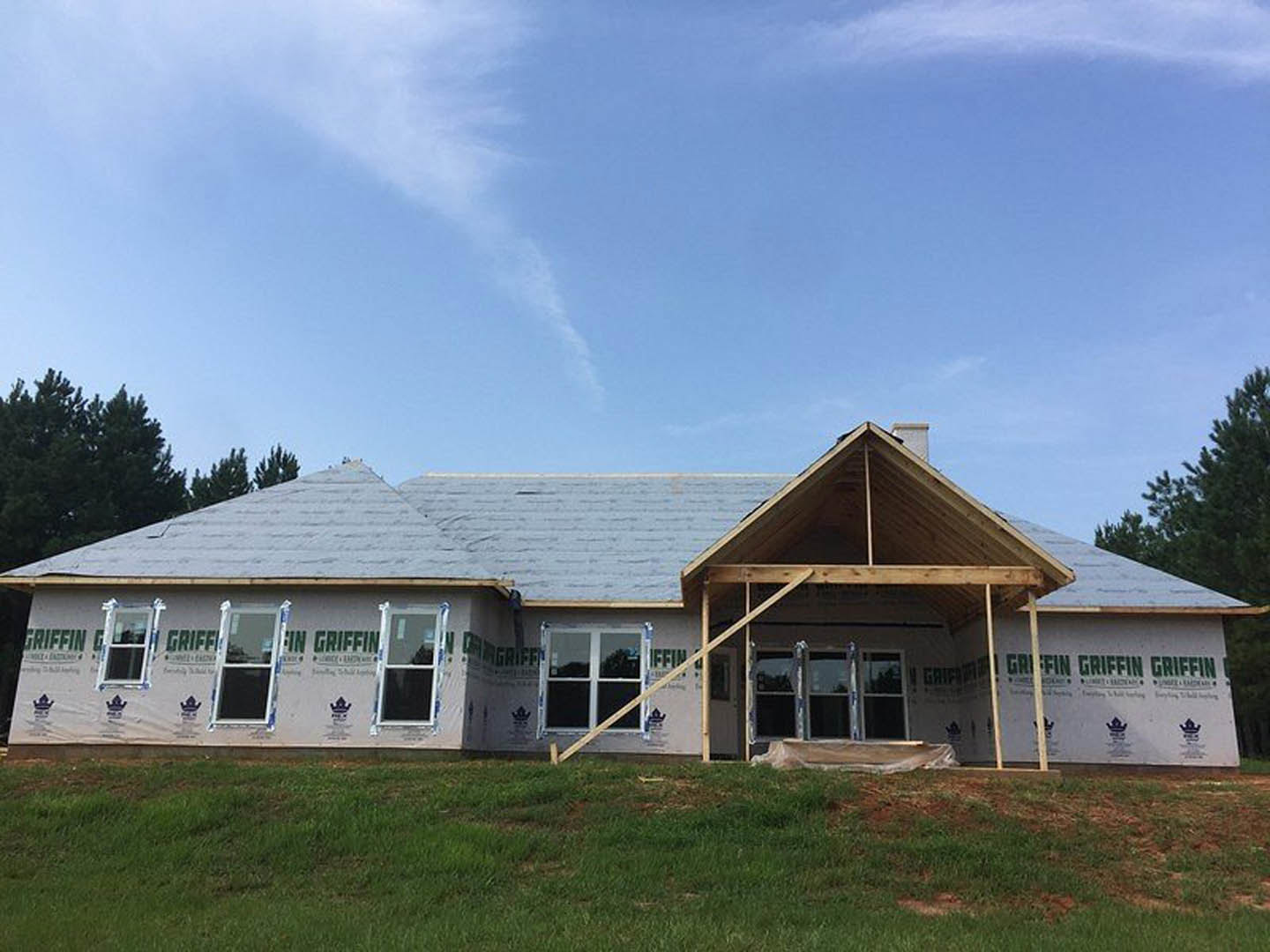 Two-story house under construction with gray shingle roof, white-framed windows, unfinished wooden porch, green grass in foreground, and blue sky with scattered clouds