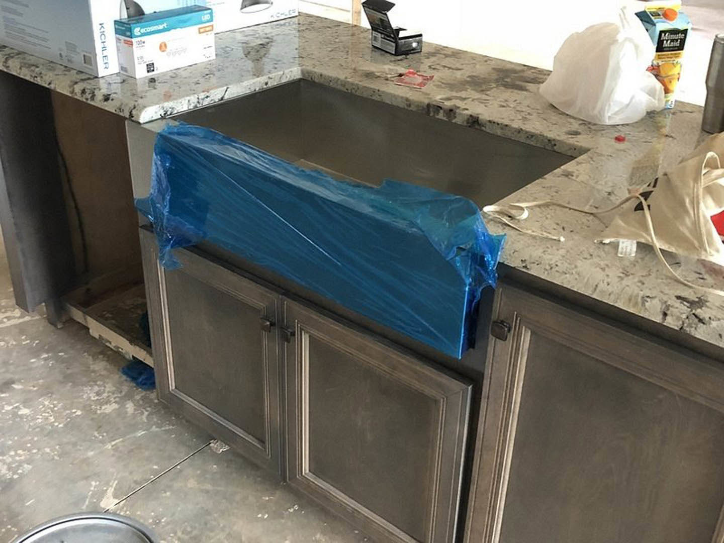 Modern kitchen with stainless steel sink, white countertop, blue plastic wrap covering a cabinet, white plastic bag on the table, and a white box with blue and white text