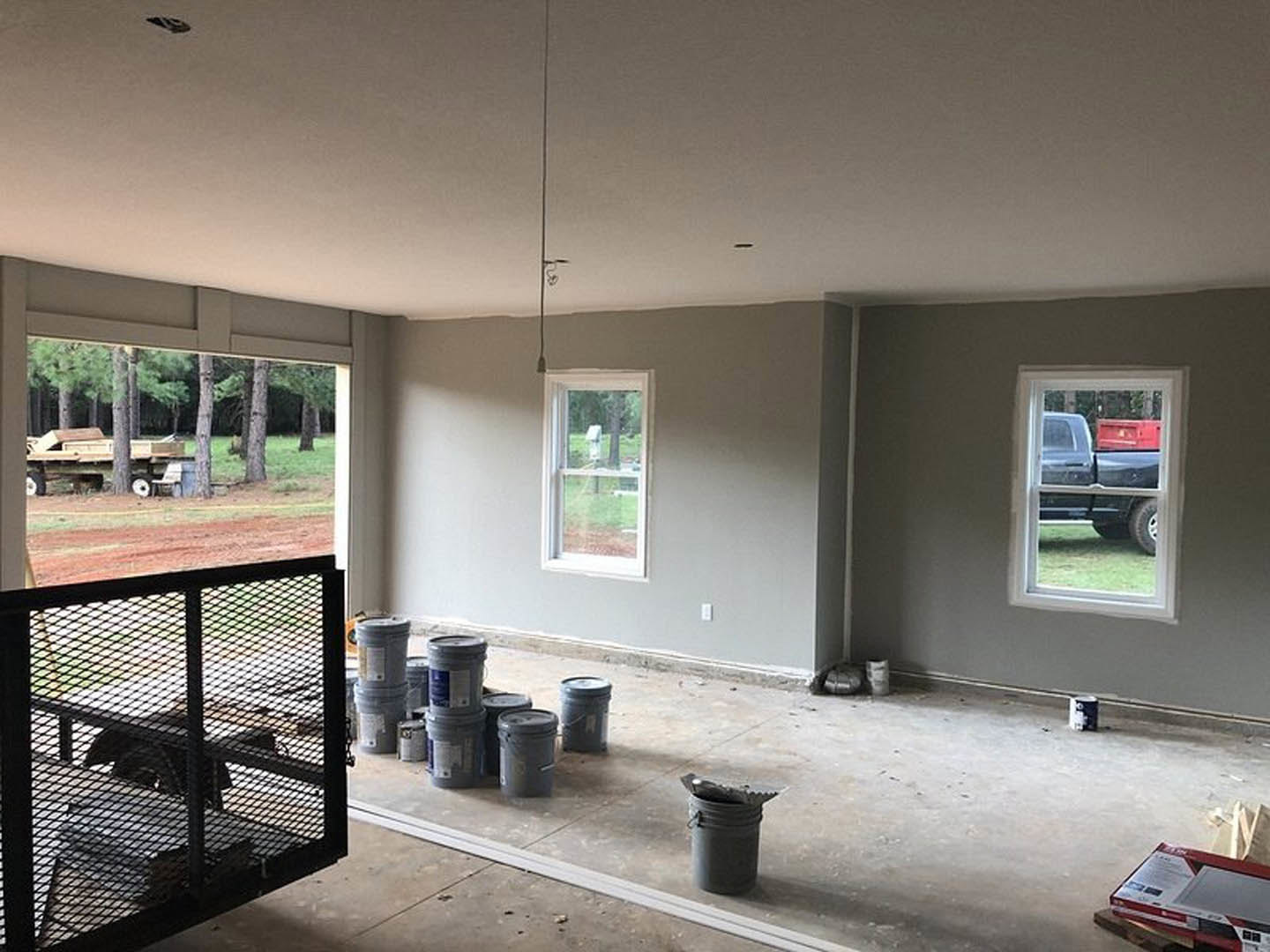 Unfinished room with paint buckets and metal containers on concrete floor, large window showing black truck parked outside, metal fence and wood visible in yard