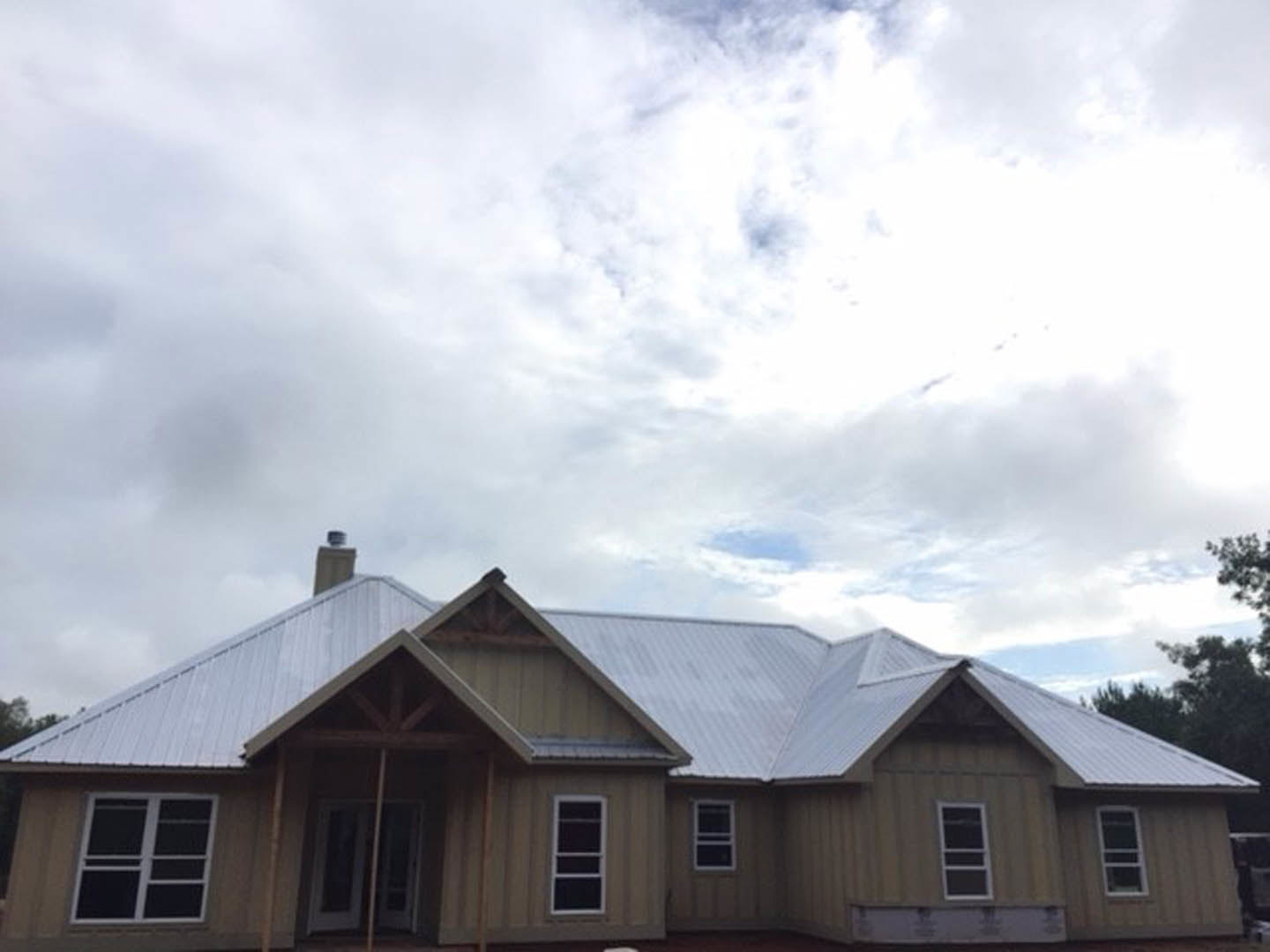 Two-story house with gray shingle roof, white-framed windows, white front door with glass panes, brick chimney, surrounded by trees under a cloudy sky