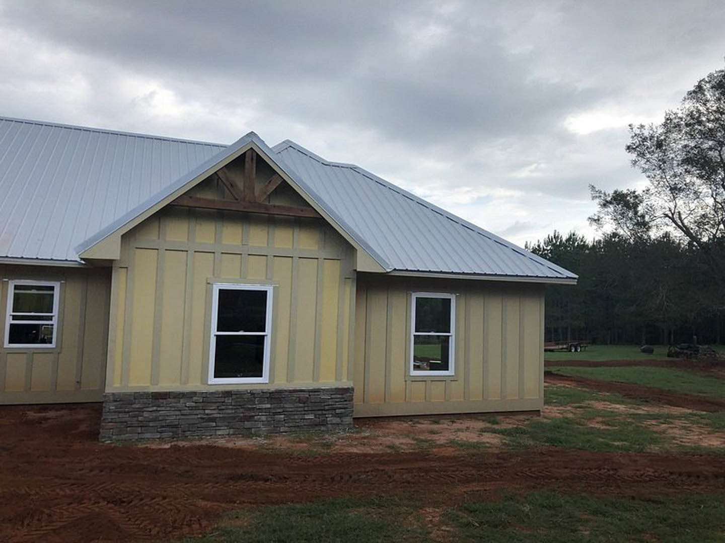 Partially built house with exposed framing, white-trimmed window, shingled roof, grassy yard, and mature tree under a cloudy sky