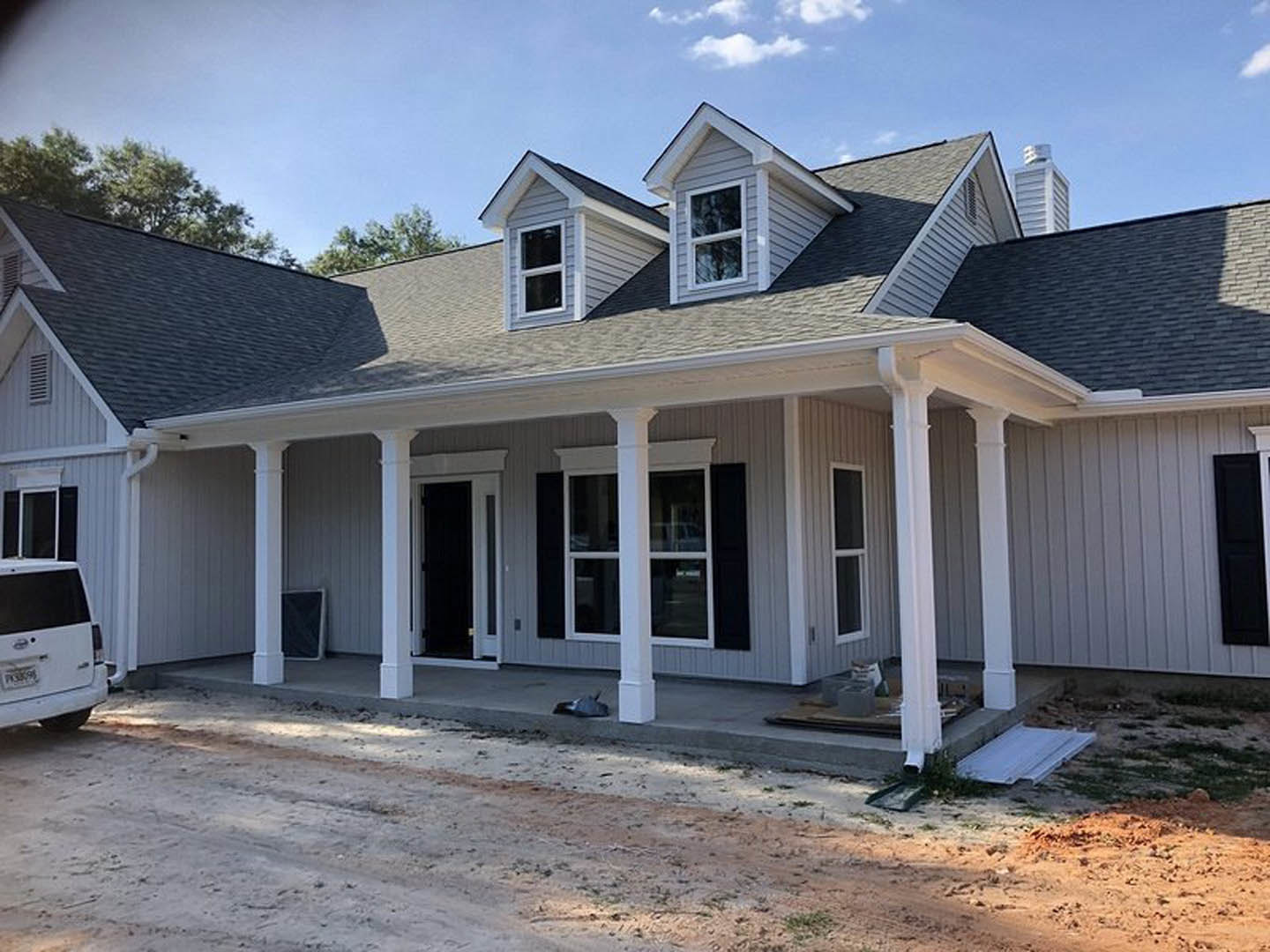 Two-story home with white siding, covered front porch supported by columns, dirt driveway, white-framed windows, and cloudy sky overhead