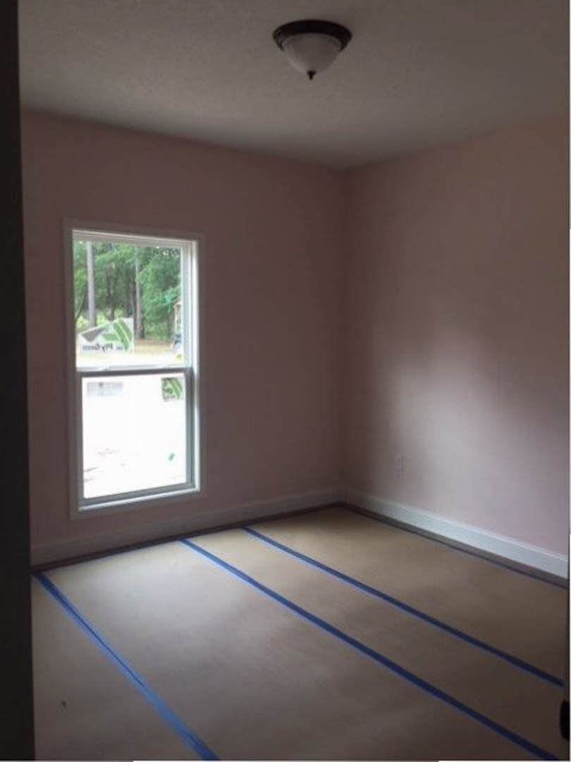 Sunlit room featuring a large window overlooking trees, wood flooring with blue stripes, white plaster walls, and a ceiling-mounted light fixture.