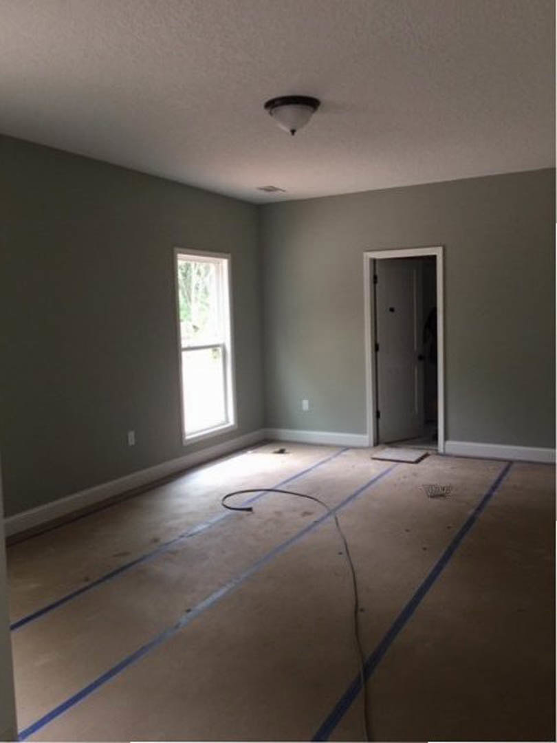 White paneled door with black hardware, white-framed window, ceiling-mounted light fixture, unfinished concrete floor marked with blue tape and visible crack