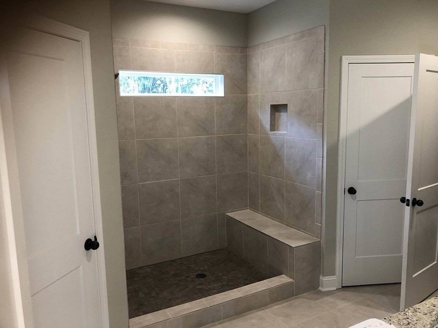 Walk-in shower with blue and white patterned tile wall, white door with black knobs, window providing natural light, and silver drain on tiled floor