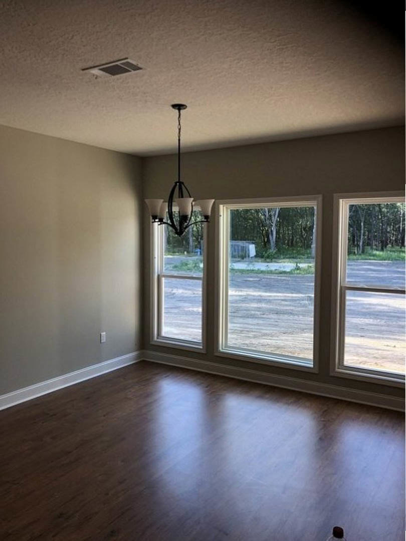 Chandelier hanging from white ceiling above hardwood floor, large windows with views of trees, grass, and road outside