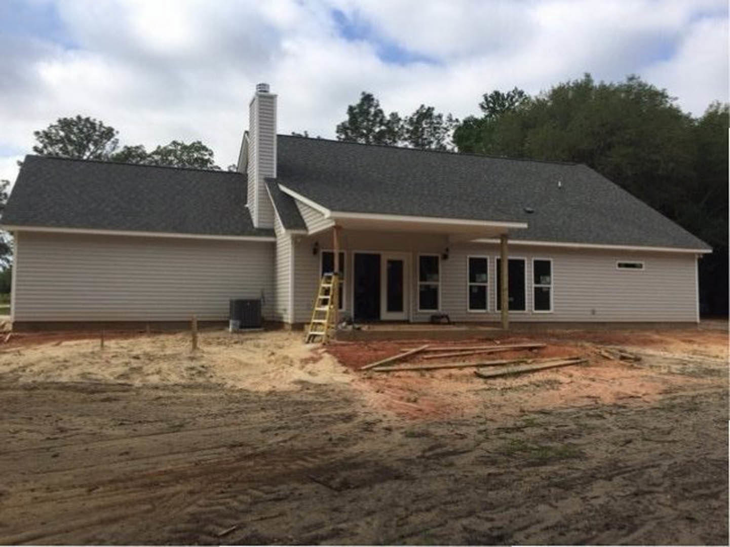 Partially built house with exposed framing, ladder leaning against exterior wall, dirt ground, white door with glass panes, black box on table, cloudy sky in background