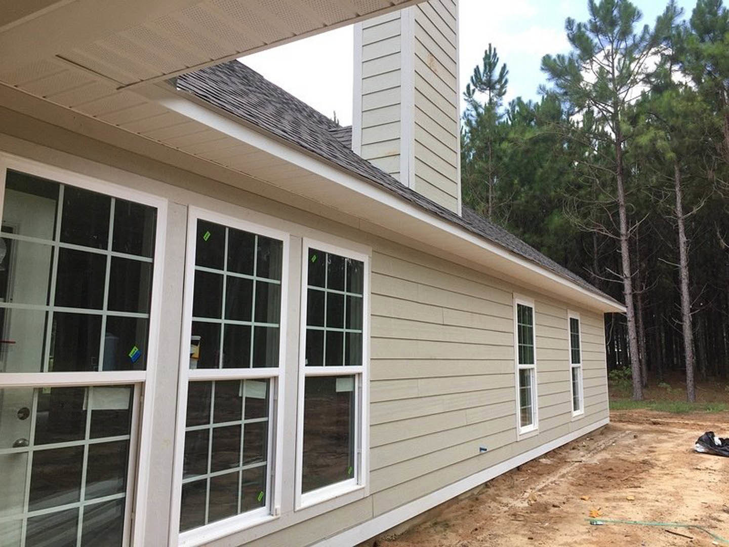 Two-story home with tall brick chimney, gray composite siding, large windows with stickers, surrounded by mature trees, construction materials visible on exterior