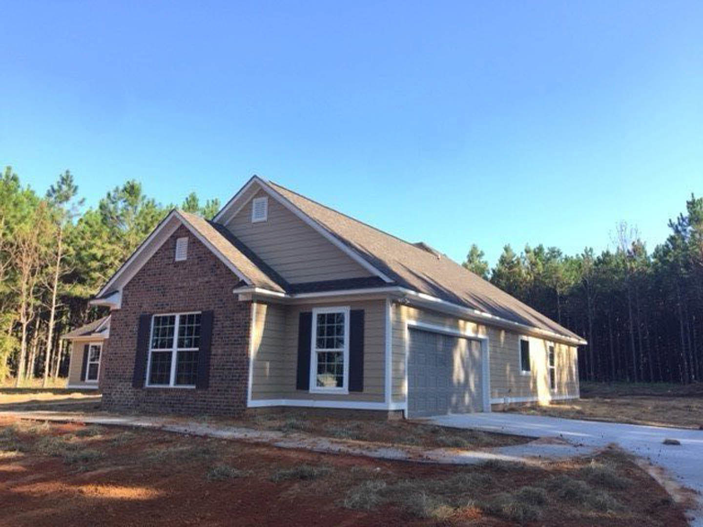 Partially built house with white multi-pane windows, unfinished garage door, dirt yard with patches of grass, surrounded by mature trees.