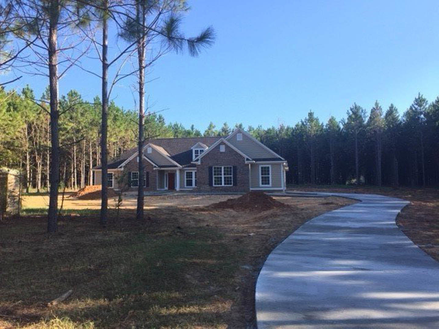 Curved concrete driveway bordered by green grass and mature trees leading to a two-story house with a red front door and white-framed windows