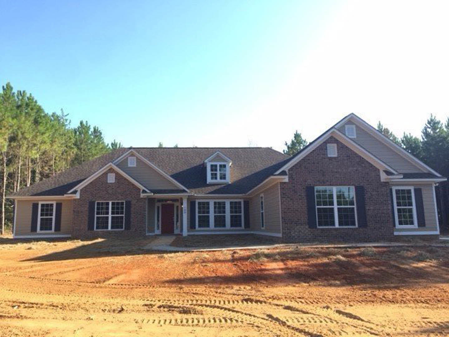 Two-story house with red front door, white-framed windows, and tire tracks across dirt yard; trees and blue sky in background.