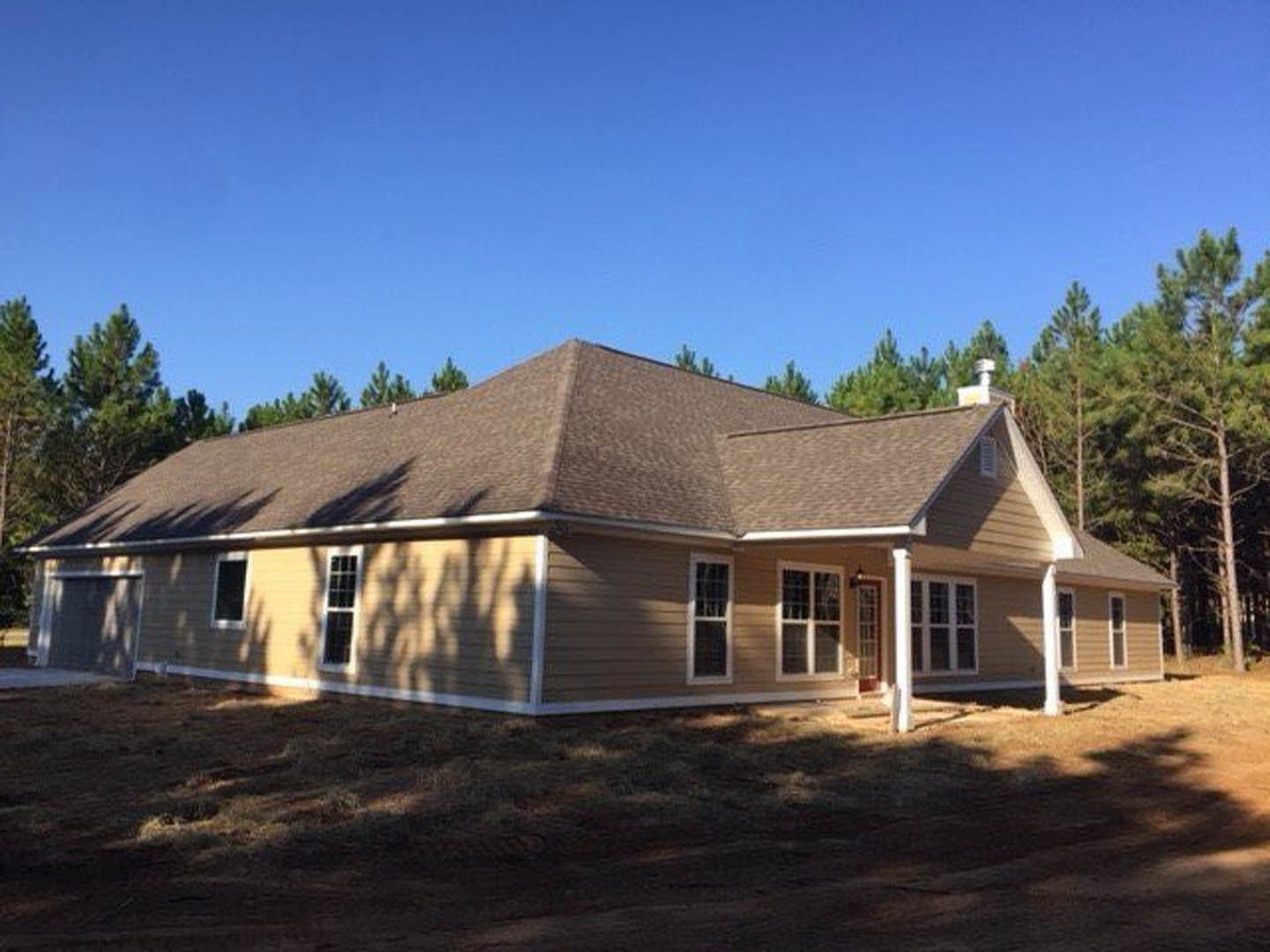 Two-story home with expansive covered porch, white roof, multiple windows with white frames, surrounded by trees under clear blue sky