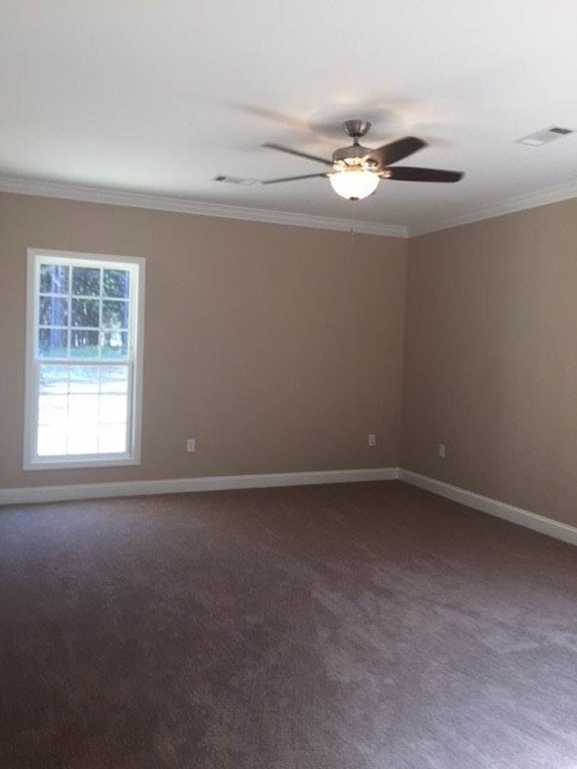 Carpeted room with white walls, ceiling fan with light fixture, large window framed in white showing leafy trees outside